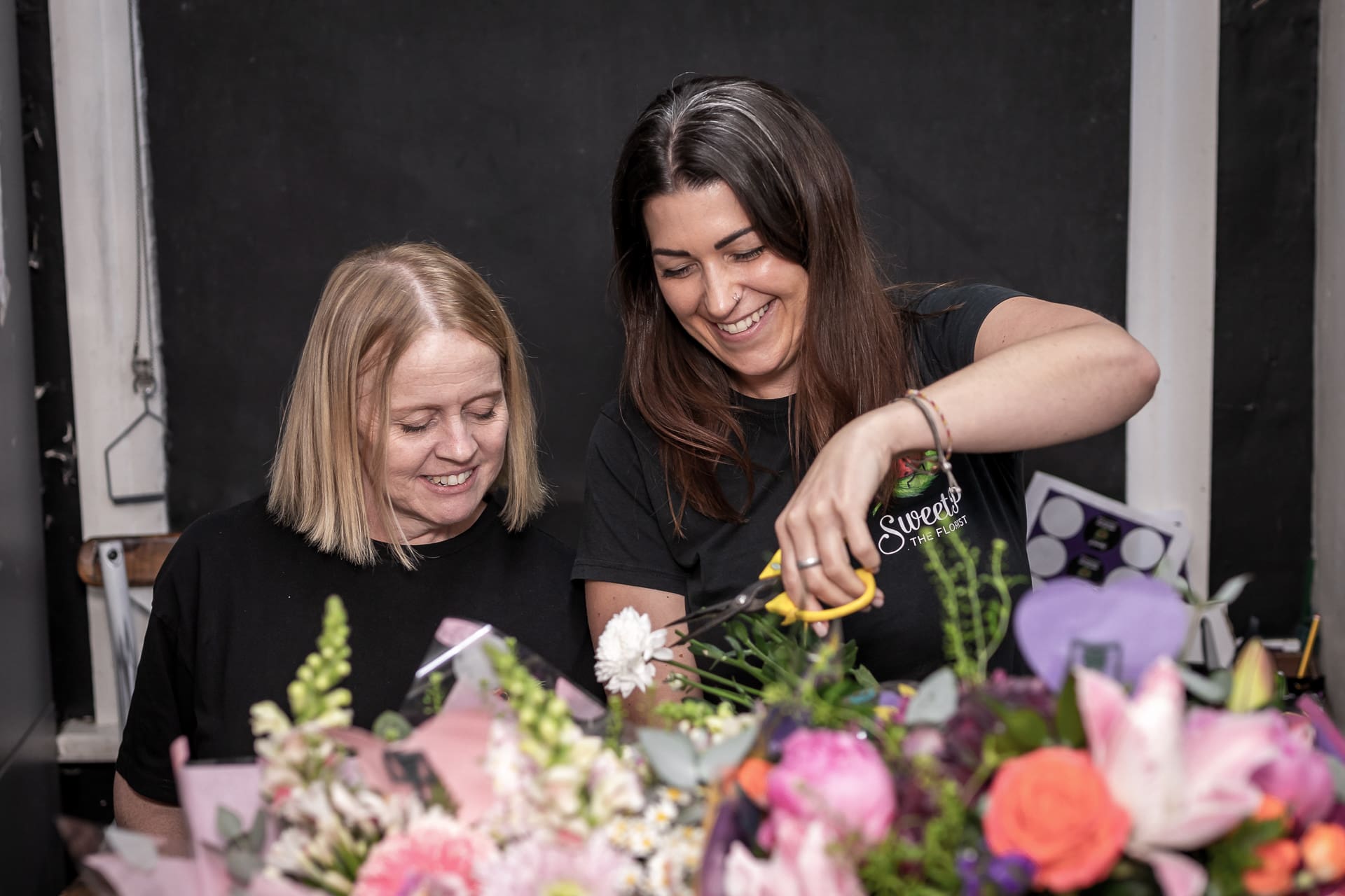Two women arranging a variety of colourful flowers together at a table, one smiling while holding scissors, with a black background behind them.