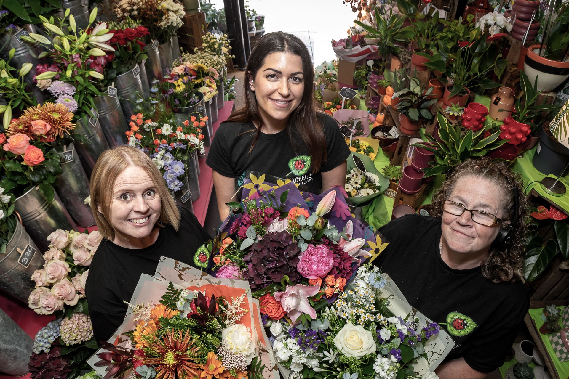 Three women in black shirts hold colourful flower bouquets and stand surrounded by various plants and floral arrangements in a flower shop.