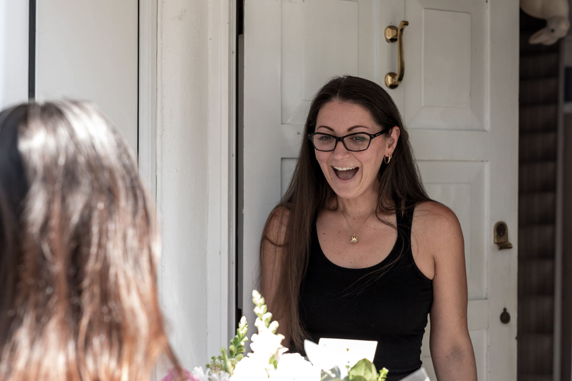 A woman with long brown hair and glasses stands at a door, smiling with surprise, whilst another person hands her a bouquet of flowers.