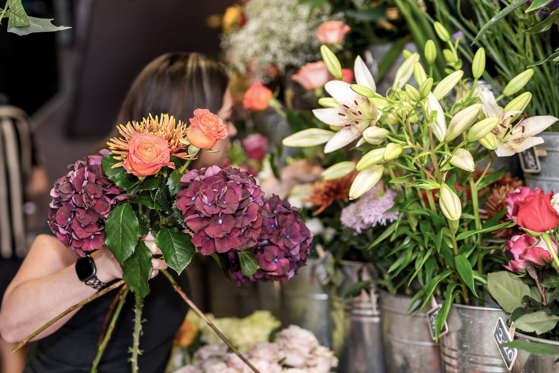 A person selecting flowers from metal buckets filled with various colourful blooms, including roses, hydrangeas, and lilies.